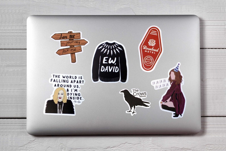May include: A collection of stickers on a laptop. The stickers include a wooden sign, a black sweater with "EW, DAVID" text, a red motel key, a woman with text, a crow, and a woman with party hat.