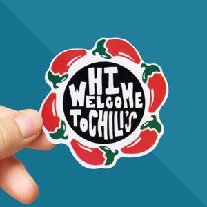 May include: A sticker with a black circle in the center, surrounded by red chili peppers. The text "HI WELCOME TO CHILI'S" is written in white, inside the black circle. The sticker is held by a hand against a teal background.
