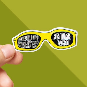 May include: A yellow sticker in the shape of sunglasses with black text. The left lens reads "Bruh Chill, I don't know why you're in a" and the right lens says "Big Time Rush". The sticker is held by a hand.