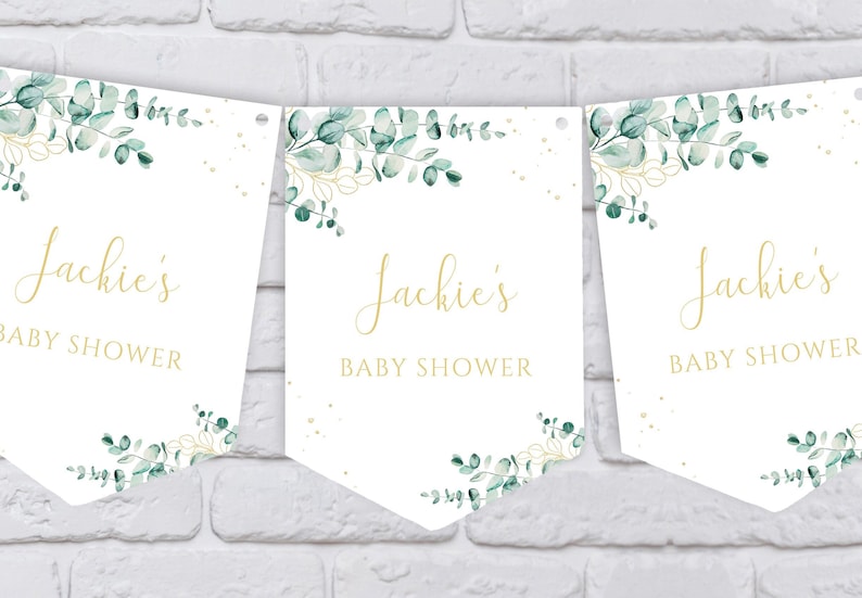 May include: Three white pennant banners with gold text and green watercolor foliage. The text on each banner reads "Jackie's Baby Shower".