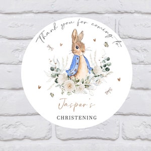 May include: Round white sticker with a thank you for coming to Jasper's christening message. The sticker features a watercolour illustration of Peter Rabbit in a blue jacket, surrounded by white flowers and greenery.