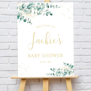 May include: White welcome sign with gold lettering and green eucalyptus leaves. The sign reads "Welcome to Jackie's Baby Shower 26-03-2023".