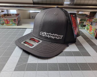 Puede incluir: Una gorra de camionero negra y gris con un logotipo "CUMMINS" blanco bordado en la parte delantera. La gorra está hecha por OTTO y tiene un parche de camionero de cinco estrellas en el lateral.