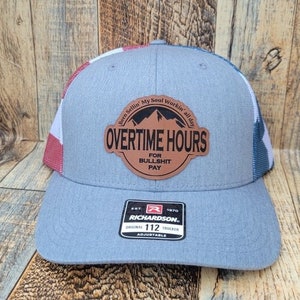 Peut inclure: Un chapeau de camionneur gris et rouge avec un patch en cuir marron qui dit "Overtime Hours for Bullshit Pay". Le patch a une silhouette de montagne et le texte "Been Sellin' My Soul Workin' all day". Le chapeau est réglable et porte le logo Richardson.