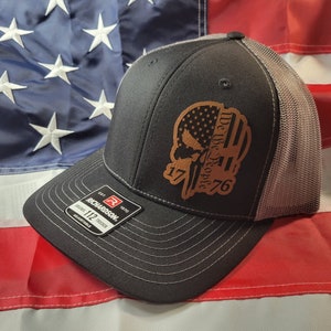 Puede incluir: Una gorra de camionero negra y gris con un parche de cuero marrón que presenta una calavera y el texto "We the People 1776". El parche tiene un diseño de bandera estadounidense roja, blanca y azul.