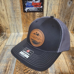 May include: Black and grey trucker hat with a leather patch showing a mountain and wave design. The hat has a curved peak and a mesh back. The patch includes the text "< > N". The hat is adjustable.