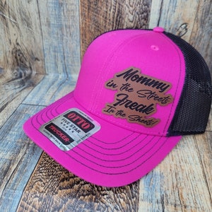 Może przedstawiać: Jaskraworóżowa czapka typu trucker z czarną siatką z tyłu. Czapka zawiera napis "Mommy in the Streets Freak in the Sheets!" na brązowej skórzanej naszywce. Czapka ma również metkę marki "OTTO".