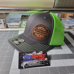 May include: A black and green trucker hat with a brown leather patch that says "Let's Keep the Dumbfuckery to a Minimum Today".
