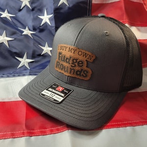 May include: A gray and black trucker hat with a brown leather patch that says "I Buy My Own Fudge Rounds".