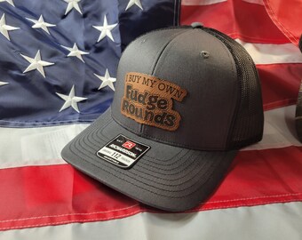 May include: A gray and black trucker hat with a brown leather patch that says "I Buy My Own Fudge Rounds".