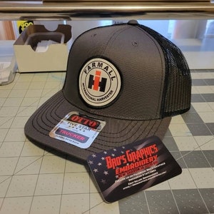 May include: A gray and black trucker hat with a red and white Farmall International Harvester logo. The hat has a black mesh back and a black and white Otto Five Star Trucker label.
