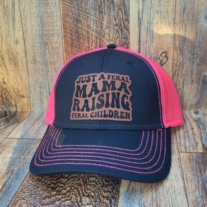 May include: A black and pink trucker hat with a brown leather patch that says "Just a Feral Mama Raising Feral Children".