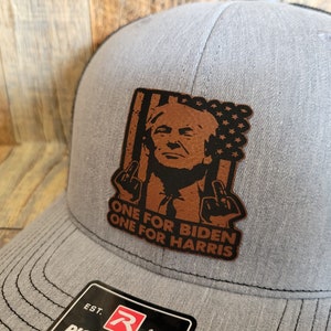 Pu&ograve; includere: Un cappello da camionista grigio con una toppa in pelle marrone che presenta una caricatura di una persona con il testo "One for Biden One for Harris".