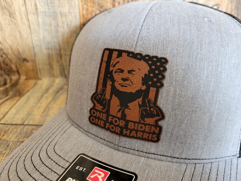Pu&ograve; includere: Un cappello da camionista grigio con una toppa in pelle marrone che presenta una caricatura di una persona con il testo "One for Biden One for Harris".