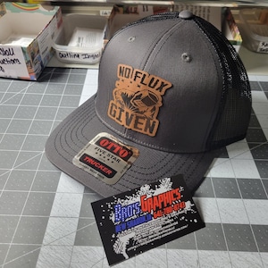 Può includere: Un cappellino da camionista grigio con retro in rete nera e una toppa in pelle con la scritta "NO FLUX GIVEN". Il cappellino ha anche un'etichetta con la scritta "OTTO Trucker". Un biglietto da visita con il nome "BROS GRAPHICS" è davanti al cappellino.