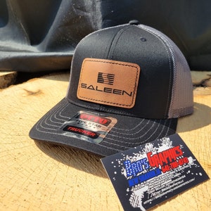 Peut inclure: Casquette de camionneur noire et grise avec un patch en cuir marron qui dit "SALEEN" en lettres rouges. La casquette a une étiquette de camionneur cinq étoiles.