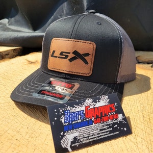 Peut inclure: Un chapeau de camionneur noir et gris avec un patch en cuir marron qui dit "LSX" en lettres noires. Le chapeau a un patch de camionneur à cinq étoiles sur le côté.