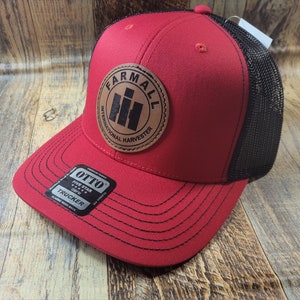 May include: Red and black trucker hat with a brown leather patch that says "Farmall International Harvester" with a black tractor logo. The hat has a black mesh back.