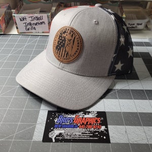 Peut inclure: Une casquette de baseball grise et noire avec un patch en cuir marron qui dit "Try That In A Small Town" avec un petit motif de drapeau américain. L'arrière de la casquette est en maille noire avec des étoiles blanches.