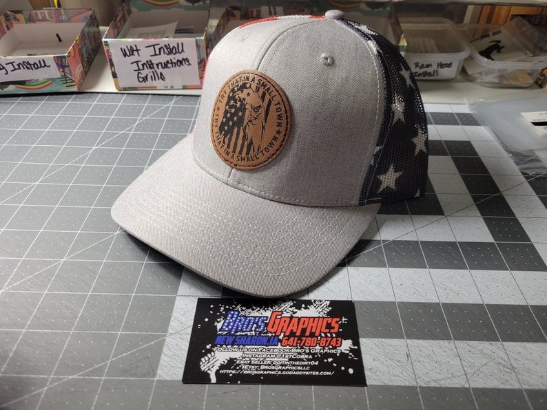Op de afbeelding: Een grijze en zwarte baseballpet met een bruin leren patch waarop "Try That In A Small Town" staat met een klein Amerikaans vlagontwerp. De achterkant van de pet is een zwart gaas met witte sterren.
