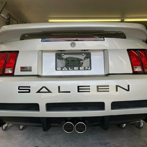 May include: A white Saleen Mustang with a black Saleen emblem on the rear bumper. The car has a black rear spoiler and dual exhaust pipes.