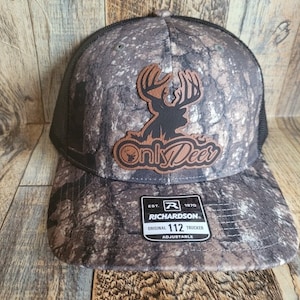 Può includere: Cappellino da camionista mimetico con toppa in pelle marrone raffigurante la sagoma di un cervo e la scritta "Only Deer". Il cappellino ha il retro in rete nera e un cinturino regolabile. L'etichetta del marchio Richardson è visibile.
