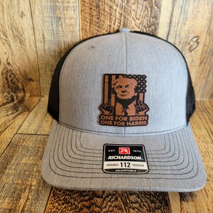 Pu&ograve; includere: Un cappello da camionista grigio e nero con una toppa in pelle marrone che presenta una caricatura di Donald Trump con il testo "ONE FOR BIDEN ONE FOR HARRIS". Il cappello &egrave; regolabile e ha un logo Richardson.