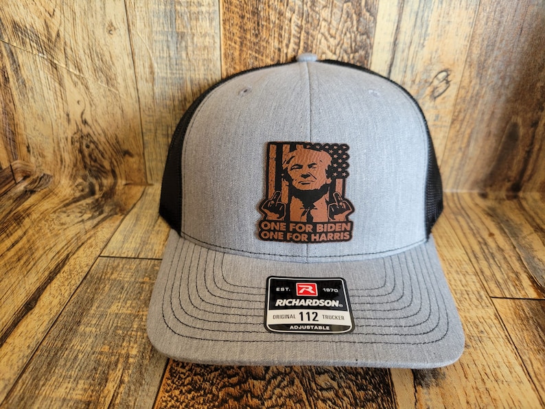 Pu&ograve; includere: Un cappello da camionista grigio e nero con una toppa in pelle marrone che presenta una caricatura di Donald Trump con il testo "ONE FOR BIDEN ONE FOR HARRIS". Il cappello &egrave; regolabile e ha un logo Richardson.