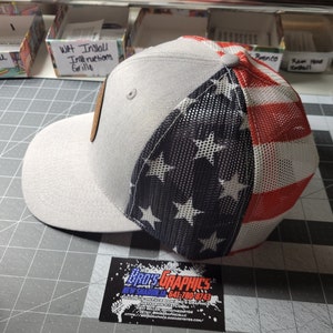 Op de afbeelding: Een grijze en witte baseballpet met een rood, wit en blauw Amerikaans vlagontwerp op de achterkant. De vlag is gemaakt van gaas.