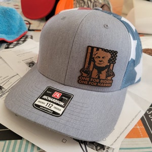 Pu&ograve; includere: Un cappello da camionista grigio e blu con una toppa in pelle marrone che presenta un ritratto di Donald Trump e il testo "ONE FOR BIDEN ONE FOR HARRIS". Il cappello &egrave; regolabile e ha un logo Richardson.