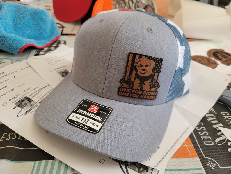 Pu&ograve; includere: Un cappello da camionista grigio e blu con una toppa in pelle marrone che presenta un ritratto di Donald Trump e il testo "ONE FOR BIDEN ONE FOR HARRIS". Il cappello &egrave; regolabile e ha un logo Richardson.