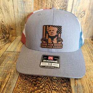 Pu&ograve; includere: Un cappello da camionista grigio e blu con una toppa di bandiera americana rosso, bianco e blu sul lato. La toppa presenta una caricatura di Donald Trump con il testo "One for Biden One for Harris". Il cappello &egrave; realizzato da Richardson e ha una cinghia regolabile di misura 112.