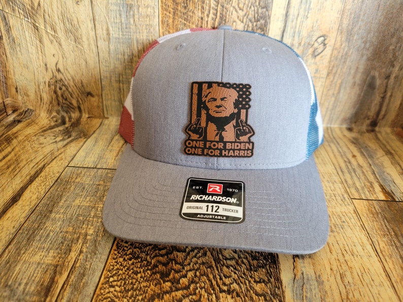 Pu&ograve; includere: Un cappello da camionista grigio e blu con una toppa di bandiera americana rosso, bianco e blu sul lato. La toppa presenta una caricatura di Donald Trump con il testo "One for Biden One for Harris". Il cappello &egrave; realizzato da Richardson e ha una cinghia regolabile di misura 112.