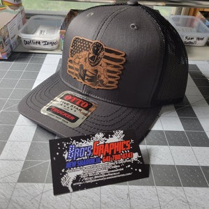 May include: A gray trucker hat with a black mesh back and a brown leather patch featuring a graphic of a person in front of an American flag. The hat has an "OTTO" tag and a business card with "BRO'S GRAPHICS" text.