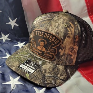 Pode incluir: Boné de caminhoneiro camuflado com parte traseira em tela preta. A frente do boné apresenta um patch com as palavras "Little Debbie Hunting Team" e a imagem de uma menina. O boné é ajustável e tem uma etiqueta Richardson.
