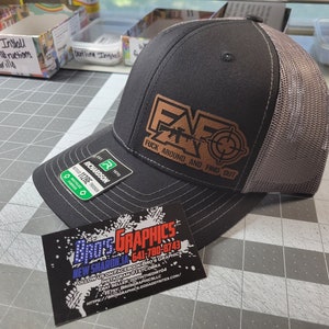 May include: A dark gray trucker hat with a brown leather patch featuring the letters "FAF", a rifle, and a target. The text "FUCK AROUND AND FIND OUT" is below the logo. The hat has a mesh back and a curved brim.