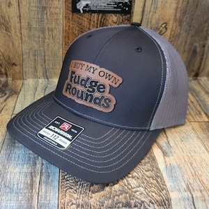 May include: A black and gray trucker hat with a brown leather patch that says "I Buy My Own Fudge Rounds".