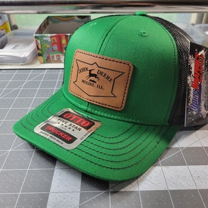 Peut inclure: Casquette de camionneur verte et noire avec un écusson en cuir marron portant le logo John Deere. La casquette a un dos en filet et une visière incurvée. L'étiquette indique "OTTO FIVE STAR TRUCKER CLASSIC MESH BACK".