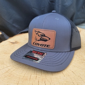Peut inclure: Un chapeau de camionneur gris et noir avec un patch en cuir marron représentant une silhouette de coyote et le mot "Coyote". Le chapeau a un dos en maille noire et un logo à cinq étoiles avec le texte "Otto Trucker".