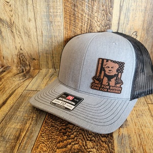 Pu&ograve; includere: Un cappello da camionista grigio e nero con una toppa in pelle marrone che presenta una caricatura di un uomo con il testo "ONE FOR BIDEN ONE FOR HARRIS".
