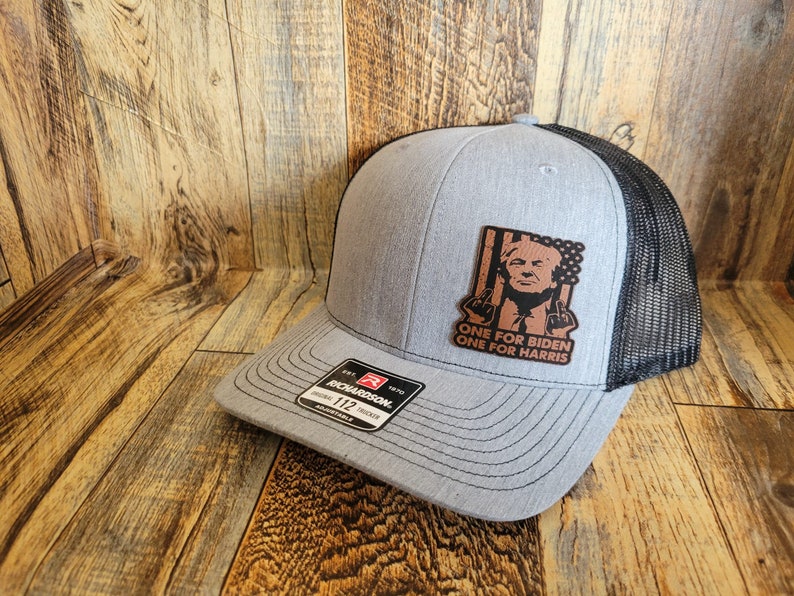 Pu&ograve; includere: Un cappello da camionista grigio e nero con una toppa in pelle marrone che presenta una caricatura di un uomo con il testo "ONE FOR BIDEN ONE FOR HARRIS".