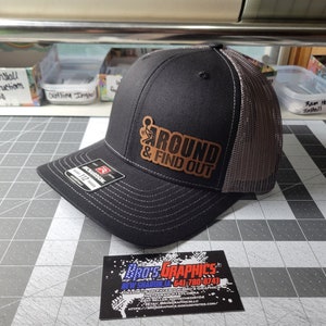 Op de afbeelding: Een zwarte en grijze truckercap met een bruin leren patch waarop "AROUND & FIND OUT" staat. De patch toont een stickfiguur met een middelvinger omhoog.