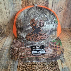 May include: A camouflage and orange baseball cap with a brown leather patch that says "Only Deer" and a deer silhouette. The cap has a black and white camouflage pattern and an orange brim. The back of the cap has a black and white label that says "Richardson 112 Original Trucker Adjustable."