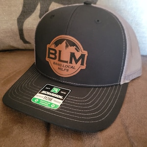 Peut inclure: Un chapeau de camionneur noir et gris avec un patch en cuir marron qui dit "BLM BANG LOCAL MILFS" et un graphique de chaîne de montagnes. Le chapeau est fabriqué par Richardson 112RE.