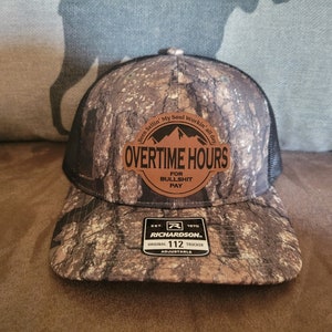 May include: A camouflage trucker hat with a brown leather patch that reads "Overtime Hours for Bullshit Pay". The hat is adjustable and has a black mesh back.