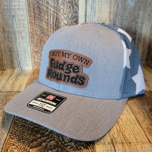 Puede incluir: Una gorra de camionero gris y azul con un patrón de estrellas blancas en la parte posterior. La parte delantera de la gorra tiene un parche de cuero marrón con el texto "I buy my own Fudge Rounds" en letras negras.