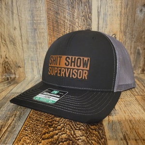 Pode incluir: Um chapéu de caminhoneiro preto e cinza com um remendo de couro marrom que diz "Shit Show Supervisor".