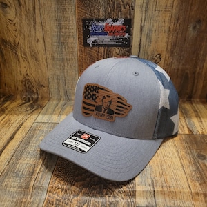 May include: A gray and blue trucker hat with a brown leather patch featuring an American flag and the text "TRUMP 2024".