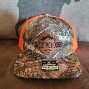 May include: A camouflage and orange trucker hat with a brown leather patch that says "Overtime Hours for Bullshit Pay". The hat is made by Richardson and is adjustable.