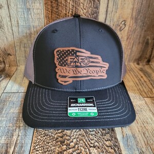 May include: A black and grey trucker hat with a brown leather patch featuring a distressed American flag with a target and the words "We The People". The hat has a black bill and a grey mesh back. The hat is made by Richardson and has a label that says "Recycled Trucker Fabrics".